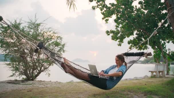 junge Freiberuflerin liegt mit Laptop in einer Hängematte am tropischen Strand - Filmmaterial, Video