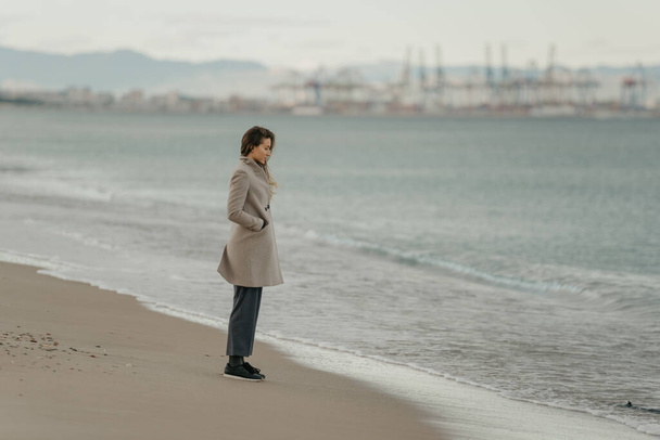 Una hermosa mujer bonita con el pelo largo en un abrigo, pantalones y zapatillas de deporte se ve en el mar
 - Foto, imagen