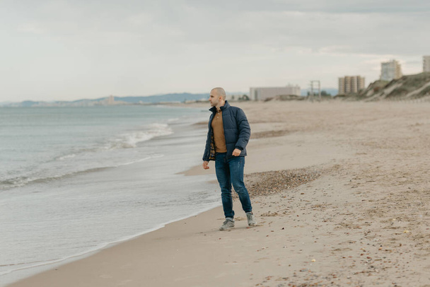 A muscular bald stylish man in the bologna jacket, jeans and sneakers poses on the shore of the Mediterranean Sea - Zdjęcie, obraz