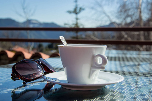 Una taza de café al aire libre, gafas de sol, primer plano de una bebida caliente y montaña en el fondo
 - Foto, Imagen