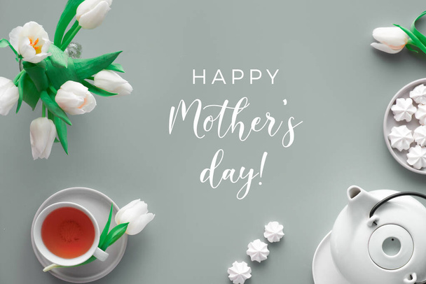 Den matek čajem a zdravím text "Šťastný den matek". Moderní moderní byt ležel, pohled shora. Čajový hrnec, sladkosti a bílé tulipány na světle šedém papíru. - Fotografie, Obrázek