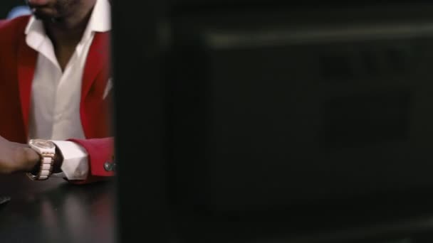 Barba matura calvo africano-americano uomo d'affari, in abito rosso, camicia bianca e occhiali eleganti utilizza smartphone tenendolo in mano, seduto in ufficio scuro con sfondo di colore blu classico. - Filmati, video