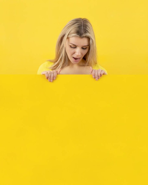 Девушка удивленно смотрит вниз на желтый лист с промо-текстом. Пространство для копирования рекламы, макета
 - Фото, изображение