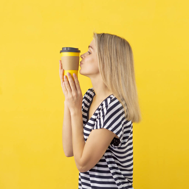 Kaffeesüchtig-Konzept. freudiges Mädchen küsst eine gelbe Einweg-Kaffeetasse auf gelbem Hintergrund - Foto, Bild