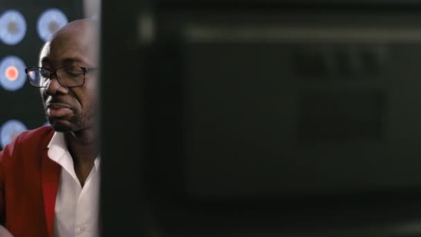 Aantrekkelijk bebaarde kale Afrikaans-Amerikaanse zakenman, in rood pak, wit shirt en stijlvolle bril kijkt naar smartphone houdt het in handen, zitten in donker kantoor met achtergrond van blauwe klassieke kleur - Video