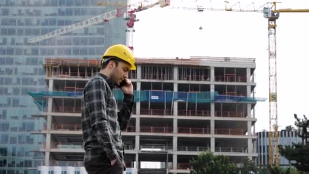 Ingenieur in een beschermende helm loopt en praat aan de telefoon op een bouwplaats. - Video