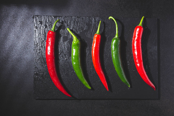 Red and green chillies on blackboard base on black background. Top view. Gastronomy and cooking condiments.  - 写真・画像