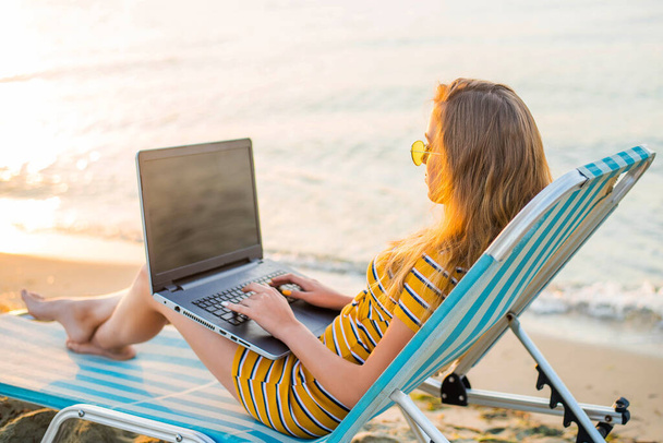 Jonge vrouw zittend op ligstoel op zee uitzicht strand met laptop past. Vrouwelijke freelance programmeur in lange chaise lounge werken codering surfen op laptop, leeg scherm. Externe werk concept. - Foto, afbeelding