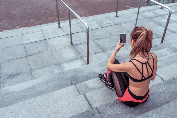 Girl athlete, in summer in city, resting after workout, holding smartphone, reading and writing message on smartphone, listening to music on headphones. Free space for copy text Fitness Motivation. - Photo, image
