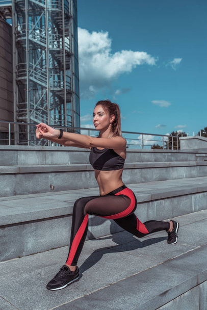 Beautiful girl, summer in city, fitness training on street, warming up muscles before training, sportswear, leggings top, sneakers. Tanned figure. Lunges and leg muscle training. - Valokuva, kuva