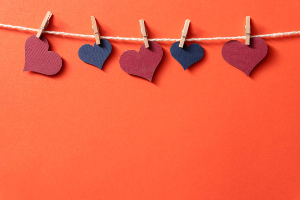 Πολύχρωμες καρδιές με σχοινί σε μικροσκοπικά μανταλάκια κρέμονται σε κόκκινο φόντο. - Φωτογραφία, εικόνα