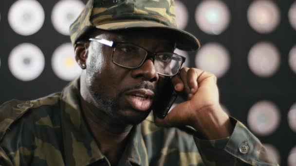 Gesicht eines bärtigen reifen schwarzen Mannes in Militärkleidung und Mütze, schwarze Brille sitzt am Schreibtisch und telefoniert in einem dunklen Raum mit verschwommenem Hintergrund. - Filmmaterial, Video