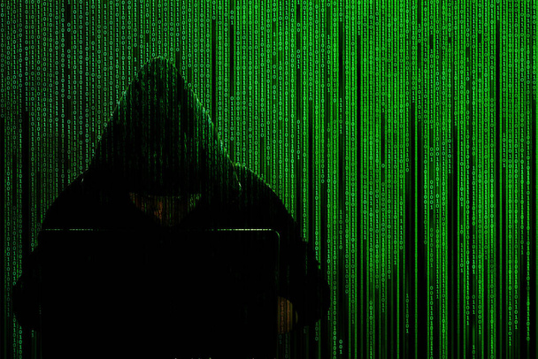 Silhouet van een hacker in een kap met een laptop op een achtergrond van een matrix van binaire computercode. Onderwerp: computerbeveiliging, hacker en computervirus, hacken database. Illustratie geïsoleerd op zwart. - Foto, afbeelding