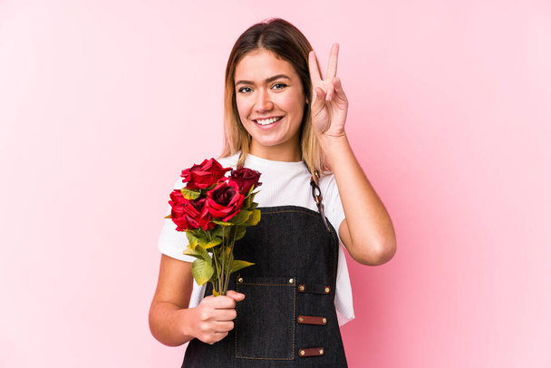 Νεαρή καυκάσια γυναίκα που κρατά τα τριαντάφυλλα απομονωμένα, δείχνοντας το σύμβολο της νίκης και χαμογελώντας πλατιά. - Φωτογραφία, εικόνα