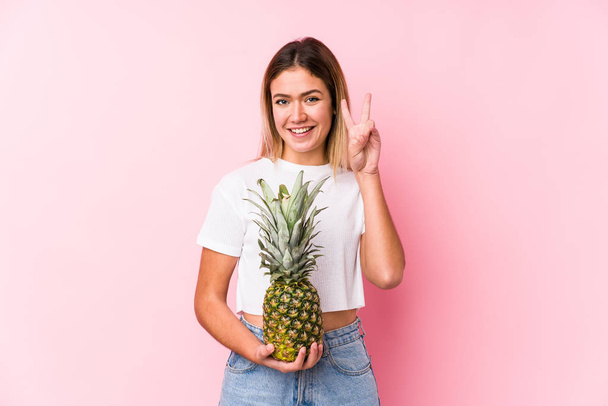 Elinde ananas tutan genç beyaz bir kadın zafer işareti gösteriyor ve gülümsüyor.. - Fotoğraf, Görsel