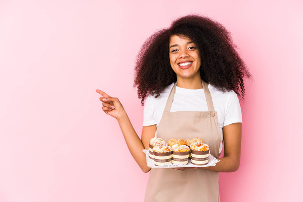 Jovem pasteleiro afro mulher segurando um cupcakes isoladoJovem padeiro afro sorrindo e apontando para o lado, mostrando algo no espaço em branco
. - Foto, Imagem