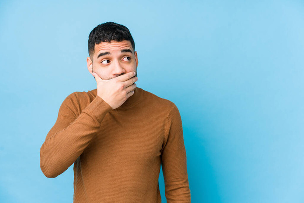 Young latin man against a blue  background isolated thoughtful looking to a copy space covering mouth with hand. - Photo, image