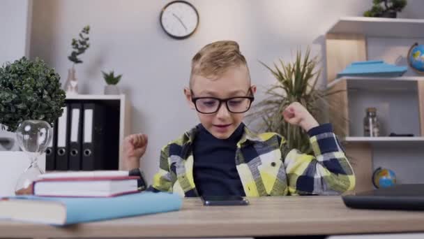 Front view of happy cheerful blond boy in glasses which raised hands up from good news on phone screen - Filmagem, Vídeo