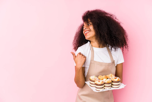 Joven pastelera afro sosteniendo una magdalenas aisladaJoven panadera afro señala con el dedo pulgar de distancia, riendo y sin preocupaciones
. - Foto, Imagen