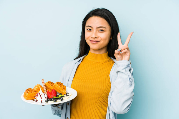 Mavi arka planda waffle tutan Çinli genç bir kadın parmakları dudaklarında bir sır saklıyor.. - Fotoğraf, Görsel