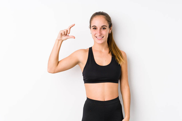 Young caucasian fitness woman posing in a white background holding something little with forefingers, smiling and confident. - 写真・画像