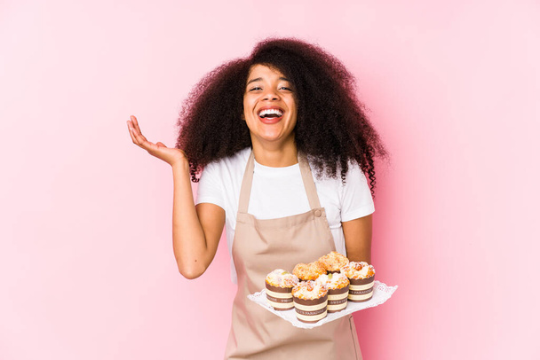 Joven pastelera afro sosteniendo una magdalena aisladaJoven panadera afro recibiendo una grata sorpresa, excitada y levantando las manos
. - Foto, Imagen