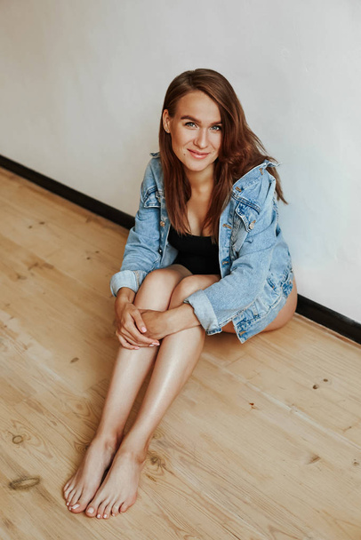 Portrait of a cute girl with a luxurious figure sitting on the floor in a stylish denim jacket. Young mother in the last month of pregnancy - 写真・画像