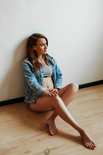 Young pregnant girl with a slim figure posing while sitting on a wooden floor. Young mom. Stylish denim jacket. - Photo, image