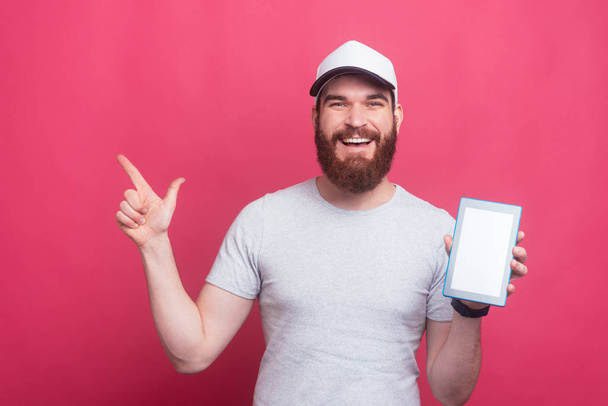 photo of joyful bearded hipster man pointing away and hodling tablet with blank screen - Fotoğraf, Görsel