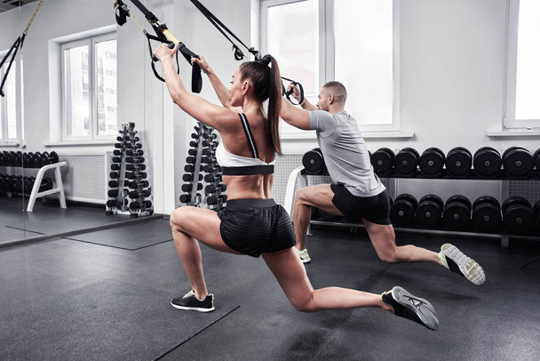 Fitness sexy brunette woman dressed top and shorts pumps arms and legs muscles with trainer in gym, personal workout program, TRX straps training - Foto, Imagem