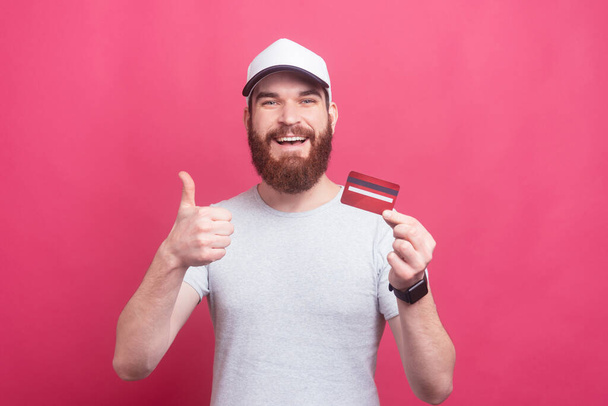 excited man with beard in t-shirt showing thumbs up and holding credit card - Foto, Bild