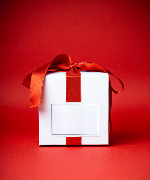 Bílá dárková krabice s prázdnou lable a červenou saténovou stuhou izolované na červeném pozadí studia - Fotografie, Obrázek