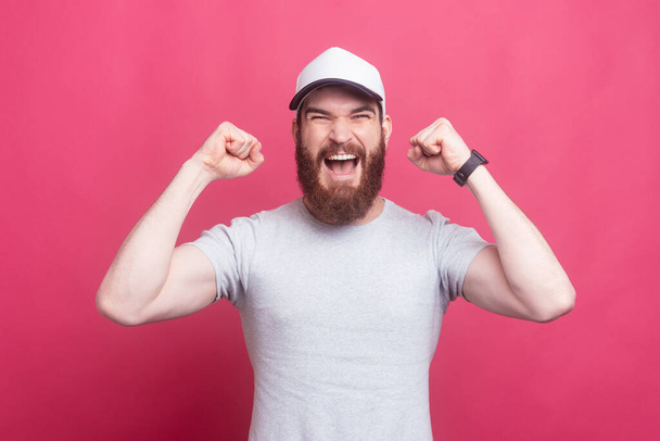 Verbazingwekkende man met baard vieren succes over roze achtergrond - Foto, afbeelding