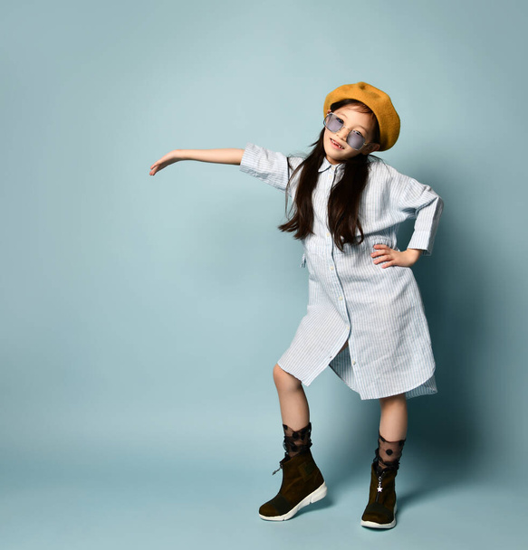 Little asian female in sunglasses, shirt dress, brown beret, boots. She smiling, showing something, posing on blue background. - Foto, Imagem