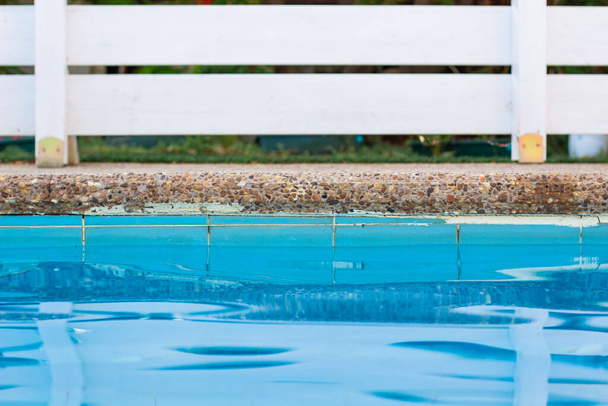 бассейн сторона мягкий фокус голубой поверхности воды передний план и белая деревянная палуба сад забор задний курорт пространство отдыха отеля
 - Фото, изображение