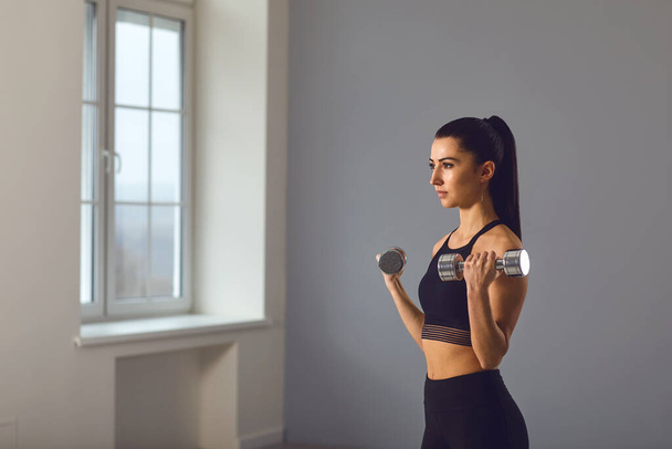 Athletic brunette girl in black sportswear with dumbbells in her hands does exercises in a room indoors - Фото, изображение