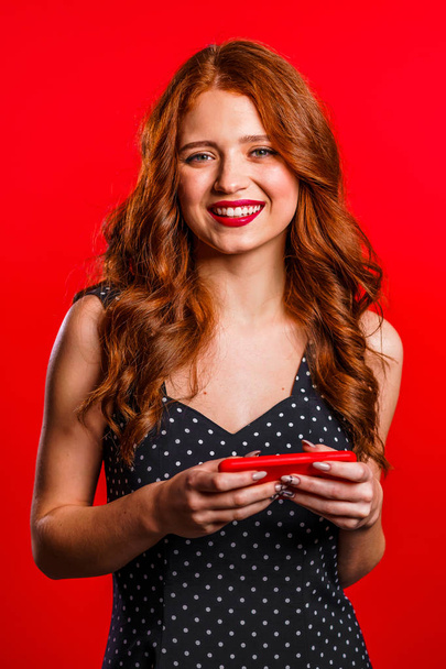 Chica con el pelo rojo utilizando el teléfono inteligente o navegar por Internet en el fondo del estudio. Tecnología moderna - aplicaciones, redes sociales
 - Foto, imagen
