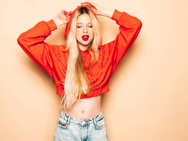 Portrait of young beautiful hipster bad girl in trendy red summer hoodie and earring in her nose.Sexy carefree smiling blond woman posing in studio.Positive model shows tongue - 写真・画像