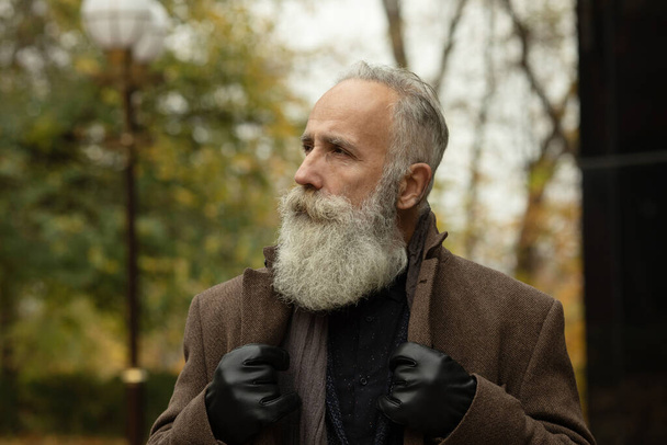 Fashionable senior man with gray hair and beard is outdoors on the street. - Фото, изображение
