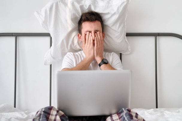 Wściekły młodzieniec w białej koszuli i piżamie trzymający białą poduszkę na głowie i laptopa na kolanach - Zdjęcie, obraz