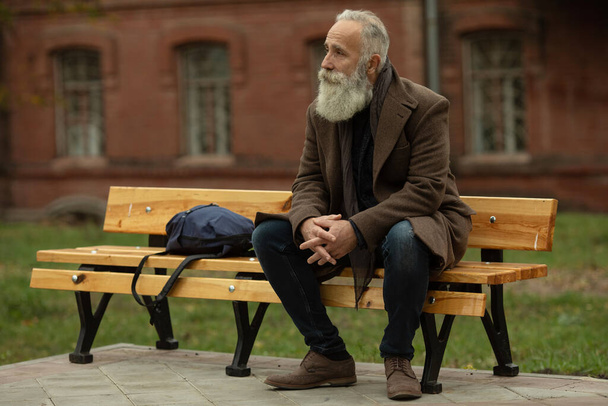 Portrait of a bearded senior man outdoors, sitting on a bench in a park. - Fotoğraf, Görsel
