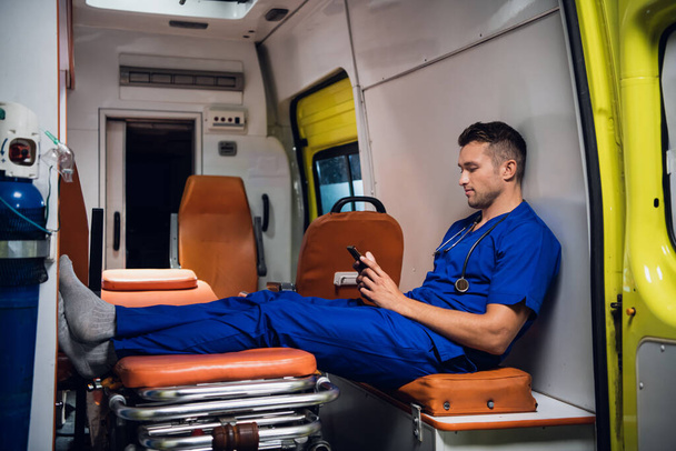 Un joven forense de uniforme se sienta con el teléfono en el coche de la ambulancia
 - Foto, imagen