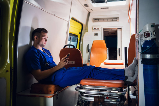 Sağlık görevlisi üniformalı genç doktor elinde telefonla ambulans arabasında oturuyor. - Fotoğraf, Görsel