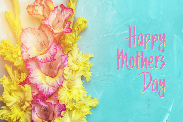 Happy Mothers Day phrase. Border frame made of yellow and pink gladiolus flowers on light blue background. Pattern of gladiolus with space for your text, holiday greeting card - Photo, Image