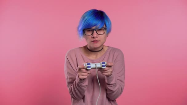 Pretty young girl with blue hairstyle playing video exciting game on Tv with joystick on pink studio wall. Using modern technology. - Felvétel, videó