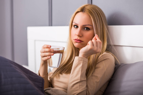 Sad woman drinking pill while sitting on bed in her bedroom. - Foto, Imagen
