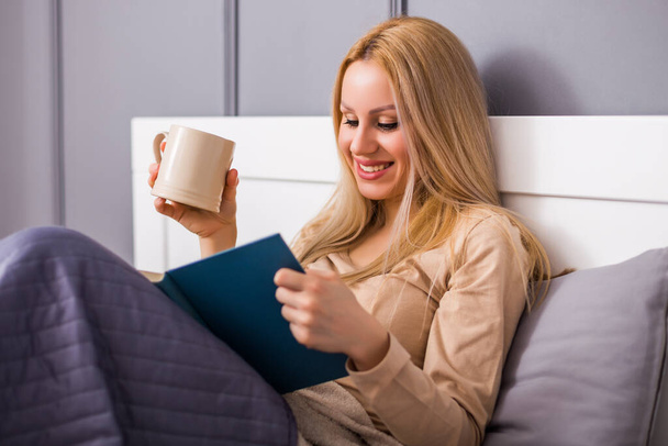 Όμορφη γυναίκα απολαμβάνει διαβάζοντας ένα βιβλίο στην κρεβατοκάμαρά της και πίνοντας τσάι ή καφέ. - Φωτογραφία, εικόνα