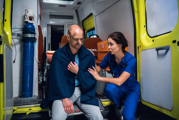 Ensihoitaja puhuu miehen kanssa ja laittaa kätensä häneen huopaan ambulanssiautossa
 - Valokuva, kuva