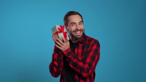 Young handsome man smiling and holding gift box on blue studio background. Guy with beard. - Metraje, vídeo