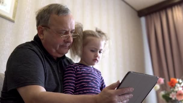 La nieta y el abuelo están sentados con una tableta. La chica frunce el ceño ante la tableta. Sentados en el sofá juntos. - Metraje, vídeo